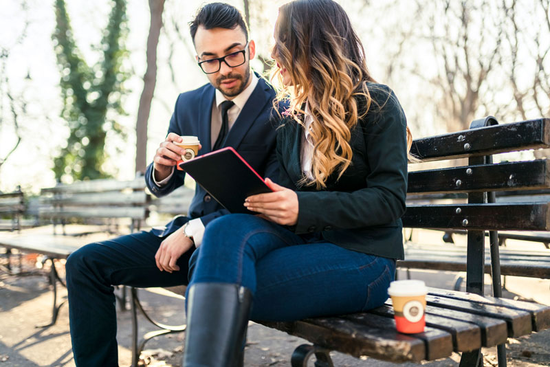 Two young professionals sit together on a park bench discussing content on an iPad