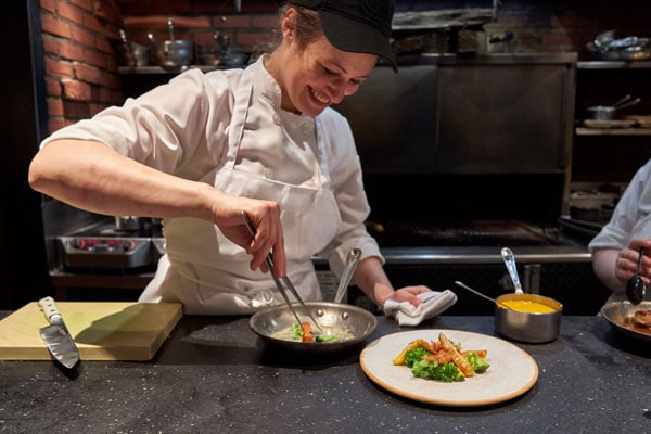A chef uses tongs to plate a vegetable dish